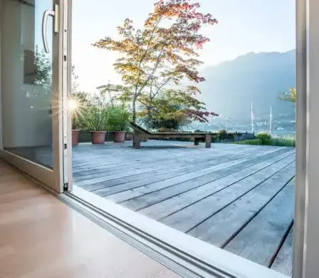 Ein Blick durch eine offene Tür auf eine Holzterrasse mit einer Liege und Bäumen.