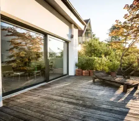 Eine Holzterrasse mit einer Bank und Topfpflanzen im hellen Sonnenlicht, in der Nähe eines modernen Hauses mit großen Glasfenstern.