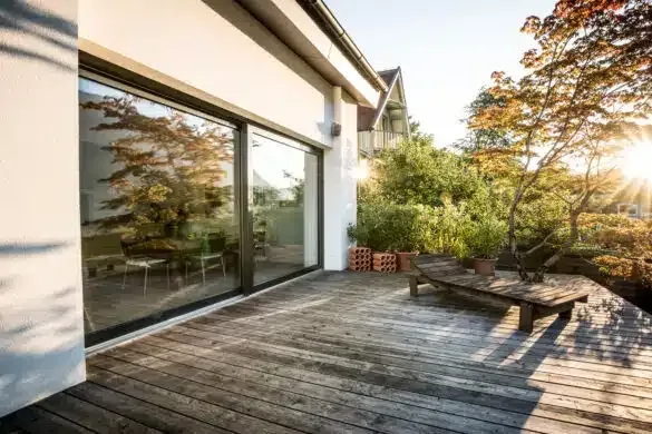 Eine Holzterrasse mit einer Bank und Topfpflanzen im hellen Sonnenlicht, in der Nähe eines modernen Hauses mit großen Glasfenstern.