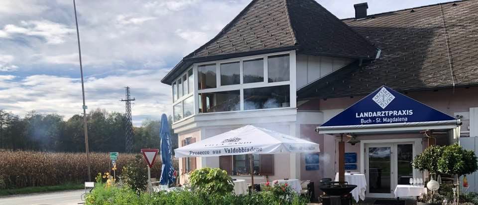 Ein Gebäude mit Glasfenstern und einem blauen Vordach mit der Aufschrift 'LANDARTZTPRAXIS' hat eine Außenbestuhlung mit einem Schirm. In der Nähe befindet sich ein Trolley mit einer Schaufel.