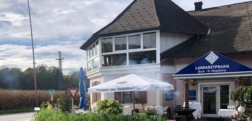 Ein Gebäude mit Glasfenstern und einem blauen Vordach mit der Aufschrift 'LANDARTZTPRAXIS' hat eine Außenbestuhlung mit einem Schirm. In der Nähe befindet sich ein Trolley mit einer Schaufel.