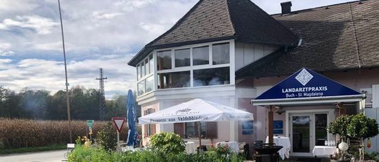 Ein Gebäude mit Glasfenstern und einem blauen Vordach mit der Aufschrift 'LANDARTZTPRAXIS' hat eine Außenbestuhlung mit einem Schirm. In der Nähe befindet sich ein Trolley mit einer Schaufel.