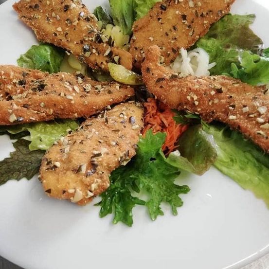 Ein weißer Teller zeigt einen Salat mit Salat, geriebenen Karotten und vier Stücken paniertem Hähnchen. Das Hähnchen ist mit Nüssen garniert.