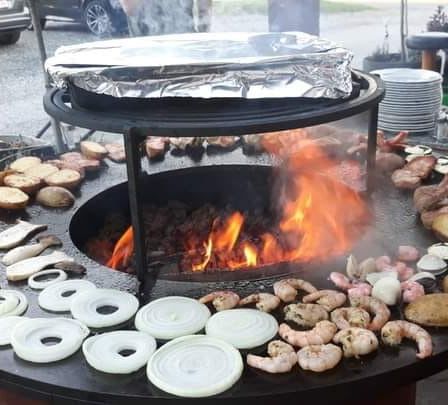 Ein großer Grill mit einem Feuerloch in der Mitte, umgeben von verschiedenen Gemüsesorten und Fleisch. Eine Person greift nach einem Stück Essen. Eine Frau mit einem Glas steht in der Nähe.