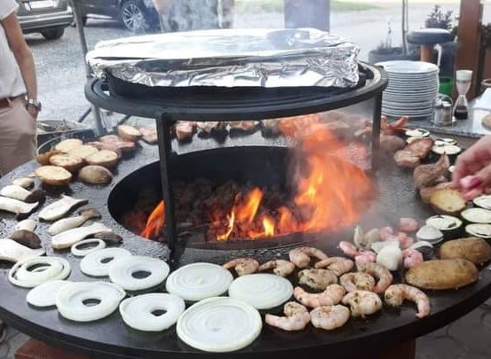 Ein großer Grill mit einem Feuerloch in der Mitte, umgeben von verschiedenen Gemüsesorten und Fleisch. Eine Person greift nach einem Stück Essen. Eine Frau mit einem Glas steht in der Nähe.