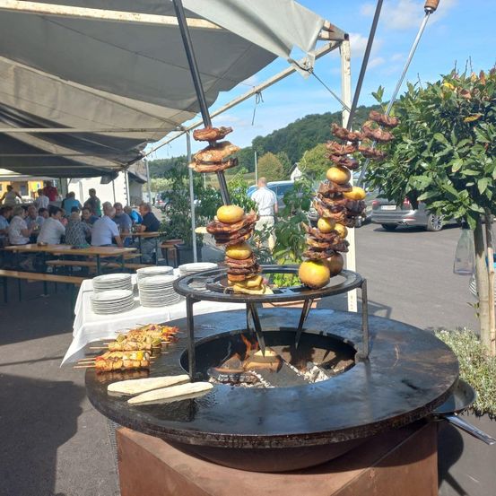 Ein Grill im Freien mit spießenden Fleisch und Früchten, unter einem Zelt, mit Leuten, die an Tischen essen, und einem geparkten Auto im Hintergrund.