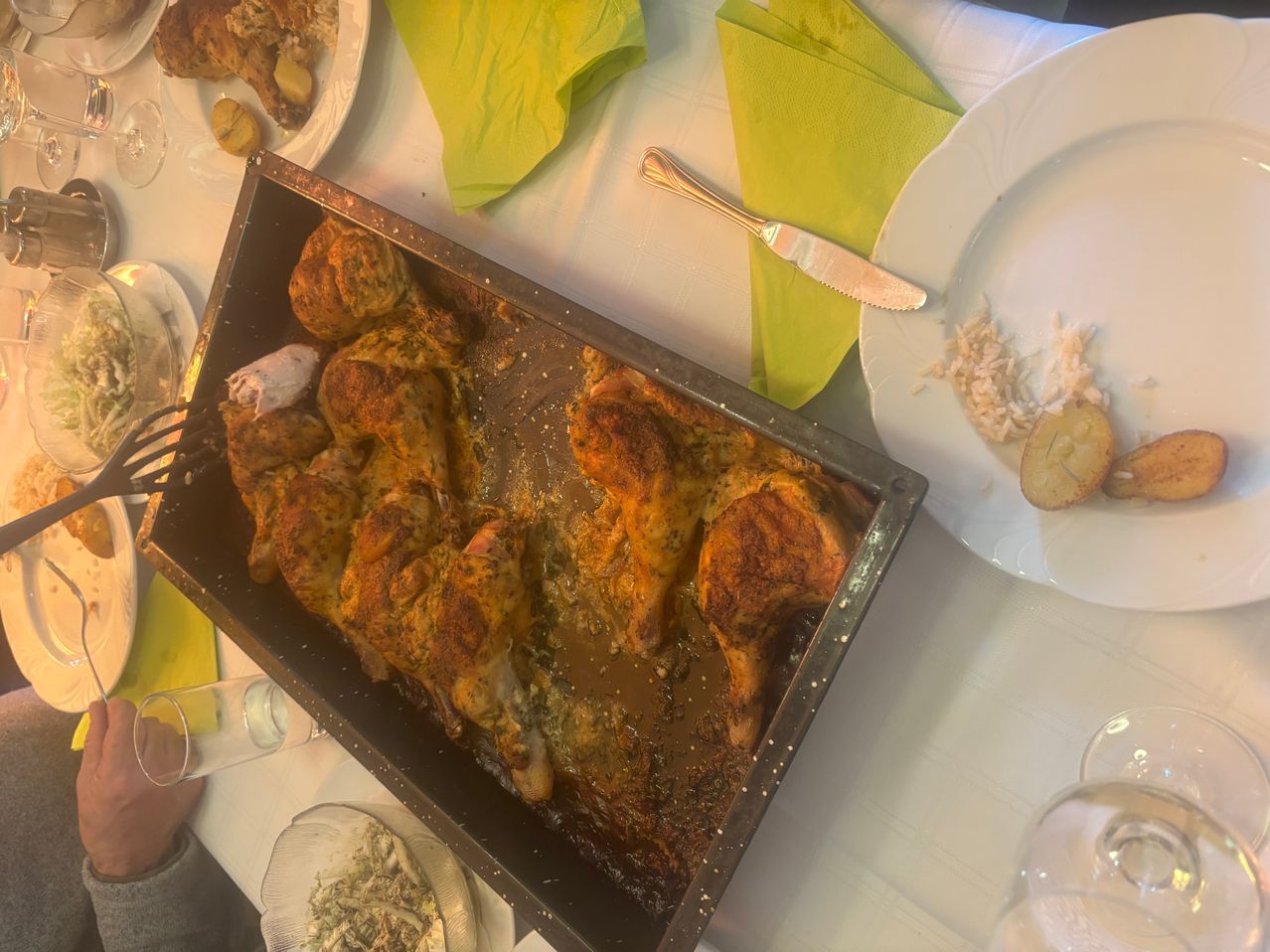 Ein Tablett mit gebratenem Huhn und Gemüse auf einem Tisch mit grünen Servietten und einem Messer. Andere Teller und ein Glas sind ebenfalls vorhanden.