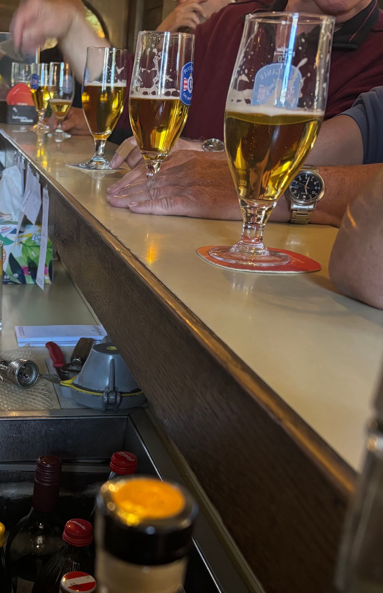 Eine Bartheke mit mehreren Gläsern Bier, eines mit Untersetzer, und die Hände einer Person auf der Theke, mit anderen Gegenständen wie einer Schachtel, einer Flasche und einer Uhr.