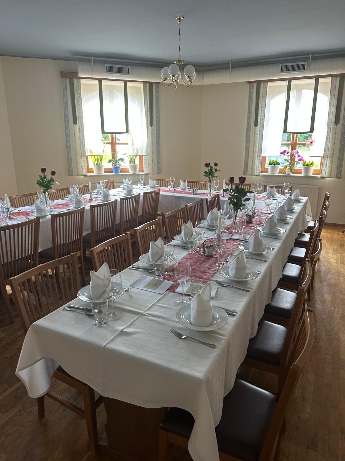 A dining room is set for a meal. Two long tables are covered with white tablecloths. Each table has multiple chairs, plates, wine glasses, and napkins. Flowers and plants are arranged on the windowsills. A chandelier hangs from the ceiling.
