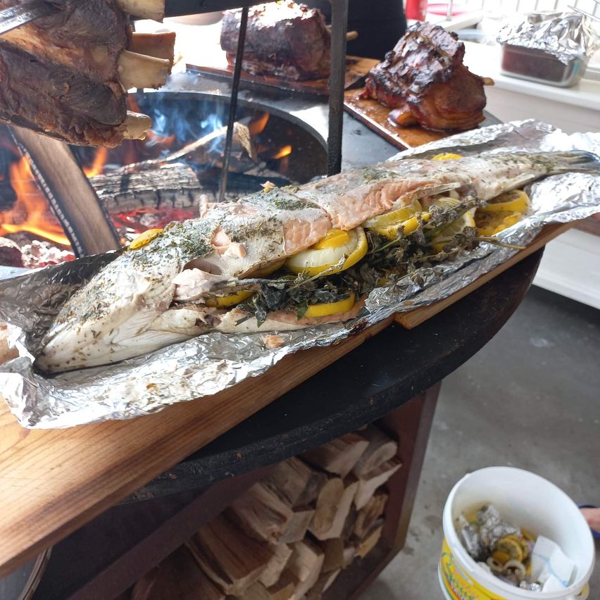 Ein Fisch, in Alufolie gewickelt und mit Kräutern und Zitronen gewürzt, ruht auf einem Holztisch mit einem Feuerkorb im Hintergrund.