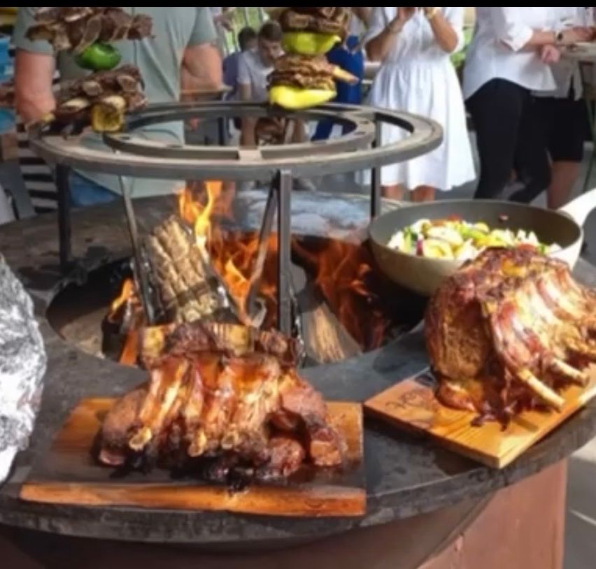 Ein runder Grill mit einem Feuerloch und Fleisch an Spießen, umgeben von einem Holztisch und einer Schüssel mit Essen. Menschen stehen herum und einige halten Teller.
