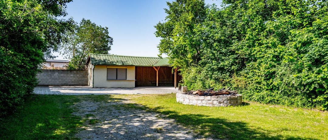 Ein kleines Haus mit grünem Dach und einem Holzschuppen ist von Bäumen und Gras umgeben. Ein Steinfeuerplatz mit Holz steht im Hof.