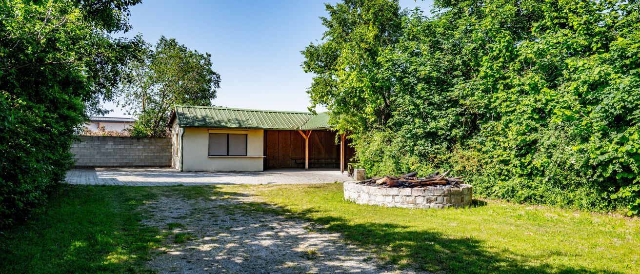 Ein kleines Haus mit grünem Dach und einem Holzschuppen ist von Bäumen und Gras umgeben. Ein Steinfeuerplatz mit Holz steht im Hof.