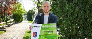 Ein Mann im Anzug hält ein KlimaTicket für die MetropolRegion Wien+BGLD und lächelt vor einem gepflasterten Weg und Grünpflanzen.