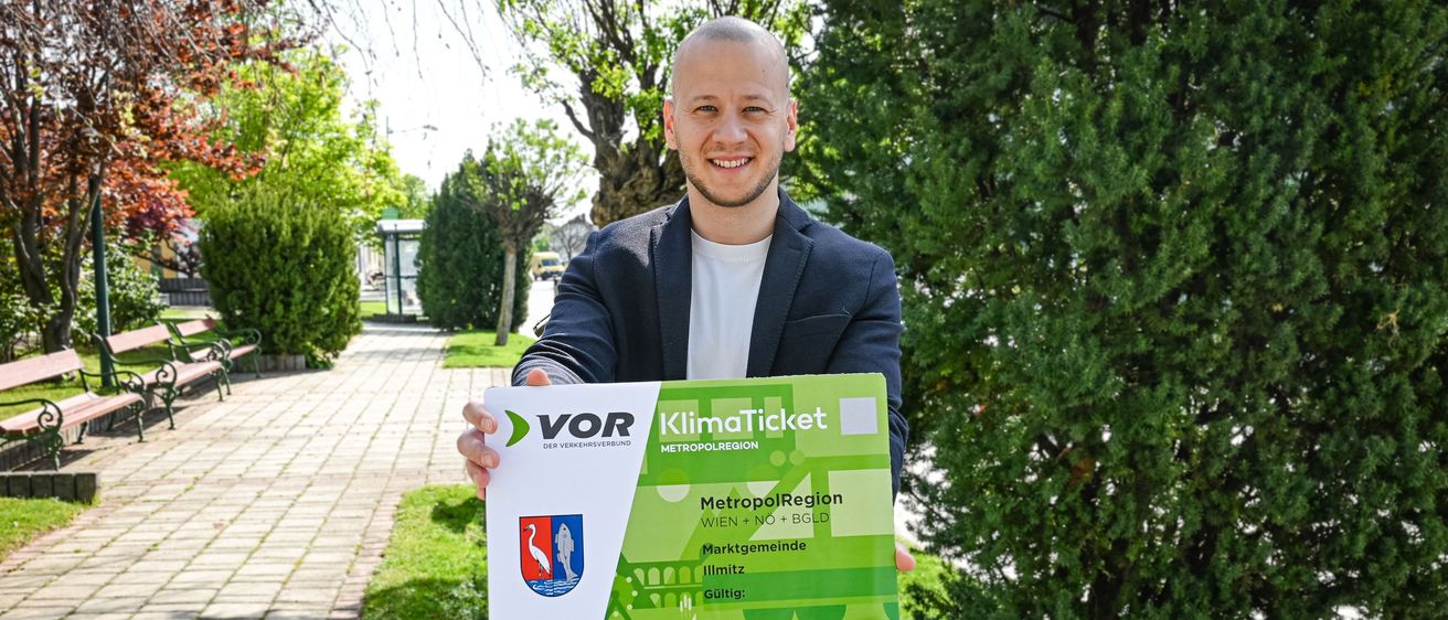 Ein Mann im Anzug hält ein KlimaTicket für die MetropolRegion Wien+BGLD und lächelt vor einem gepflasterten Weg und Grünpflanzen.