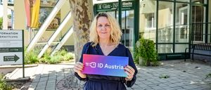 Eine Frau in einem blauen Kleid hält ein Schild mit der Aufschrift ID Austria vor einem Gebäude mit einer Glaswand.