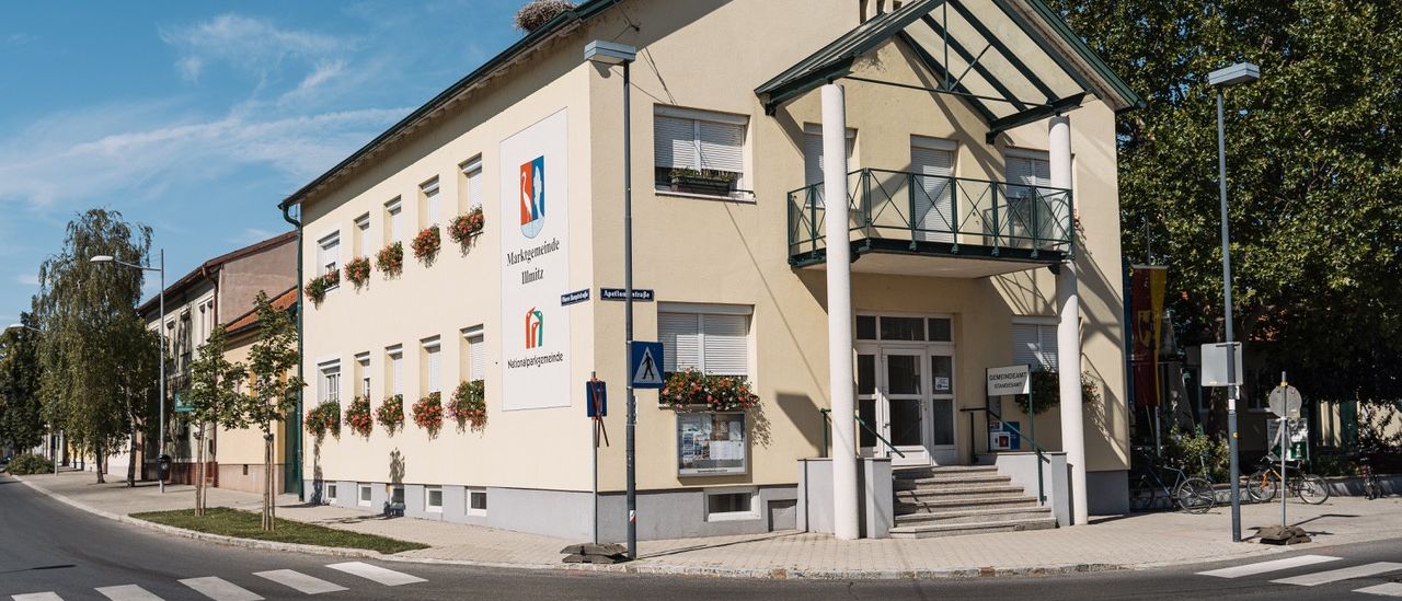 Ein beigefarbenes Gebäude mit mehreren Fenstern und Topfpflanzen steht an der Ecke. Es verfügt über eine verglaste Veranda und eine Treppe, die zum Eingang führt. Ein Schild und ein Nest befinden sich auf dem Dach.