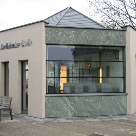 Ein Gebäude mit einem Schild, das 'Burthohmanns-Quell' steht, mit modernem Design, Glasfenstern und einer grünen Marmorwand. Eine Bank befindet sich auf der linken Seite und ein Auto ist draußen geparkt.