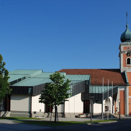 Ein Gebäude mit einem grünen Dach und einem Turm mit einer Uhr und einer Kuppel ist an einem sonnigen Tag. Vor dem Gebäude steht ein Baum.