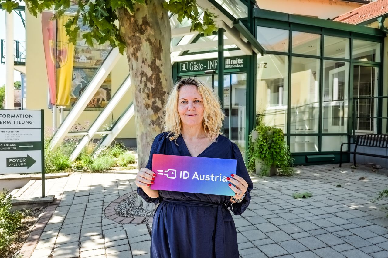 Eine Frau in einem blauen Kleid hält ein Schild mit der Aufschrift ID Austria vor einem Gebäude mit einer Glaswand.