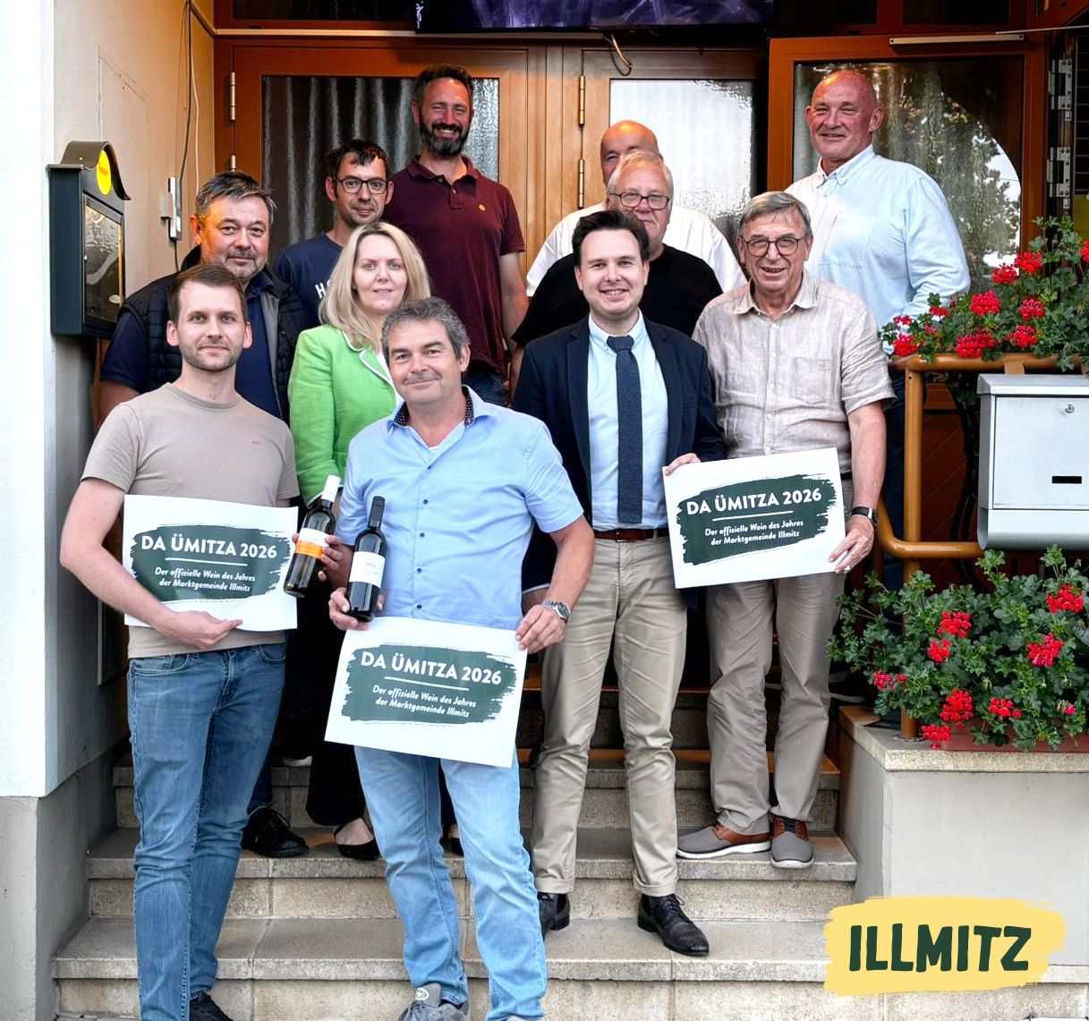 Eine Gruppe von Menschen posiert für ein Foto mit Weinflaschen und Schildern vor einem Gebäude. Die Schilder lesen DA UMITZA 2026.