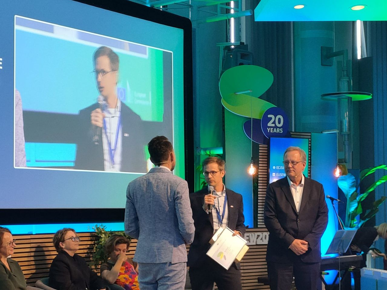 Ein Konferenzraum mit einer großen Leinwand, auf der ein sprechender Mann abgebildet ist. Drei Männer stehen vor der Leinwand, einer hält ein Mikrofon und ein Papier. Andere sitzen im Hintergrund.