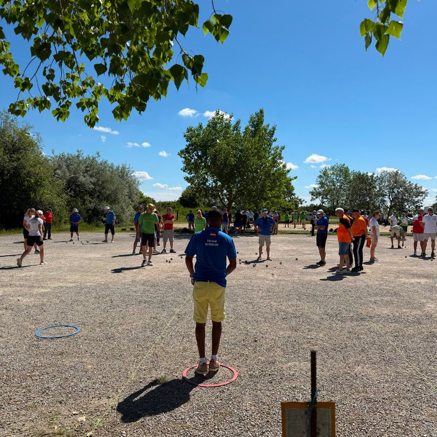 Eine Gruppe von Menschen spielt draußen auf einem Kiesplatz ein Spiel. Einige stehen in Kreisen, während andere herumlaufen. Der Himmel ist klar mit einigen Wolken.
