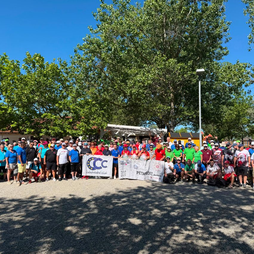Eine große Gruppe von Menschen in verschiedenen farbigen Shirts und Mützen posiert für ein Foto unter blauem Himmel mit Bäumen im Hintergrund. Einige halten Banner mit Text in den Händen.