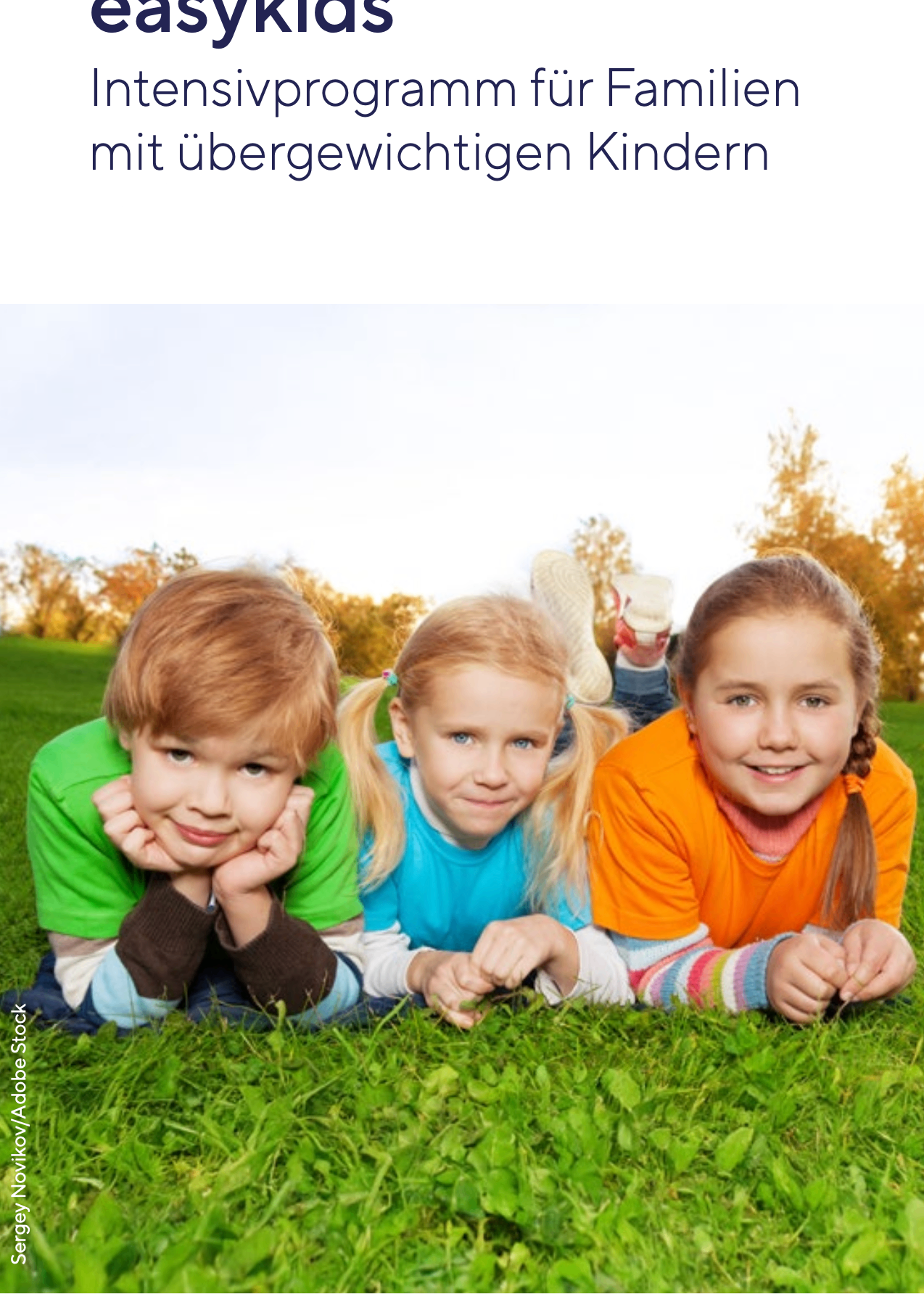 Drei Kinder liegen auf einem Grasfeld und lächeln in die Kamera. Der Junge links trägt ein grünes Shirt, das Mädchen in der Mitte ein blaues Shirt und das Mädchen rechts ein orangefarbenes Shirt. Hinter ihnen sind Bäume mit gelben Blättern zu sehen.