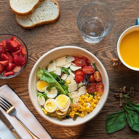 Ein Holztisch hat einen Salatschüssel mit Eiern, Tomaten und Mais, ein Glas Wasser, eine Suppenschüssel und einen Teller mit Erdbeeren.