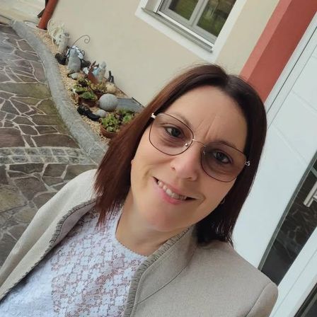 Eine Frau mit Brille und einer grauen Jacke lächelt vor einem Haus mit Fenster und einem Garten mit verschiedenen Pflanzen.