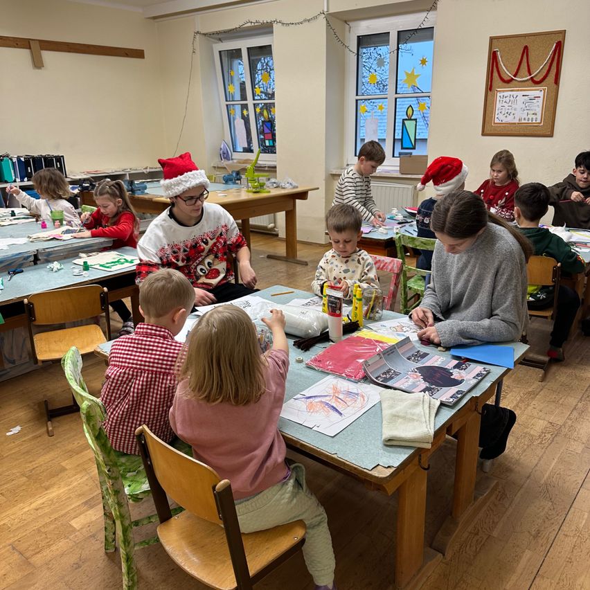 Ein Klassenzimmer voller Kinder und Erwachsener. Einige Kinder sitzen an Tischen und arbeiten an Bastelarbeiten, während andere stehen und eine Frau betrachten, die sie anleitet. Der Raum hat weiße Wände und Fenster mit Dekorationen.