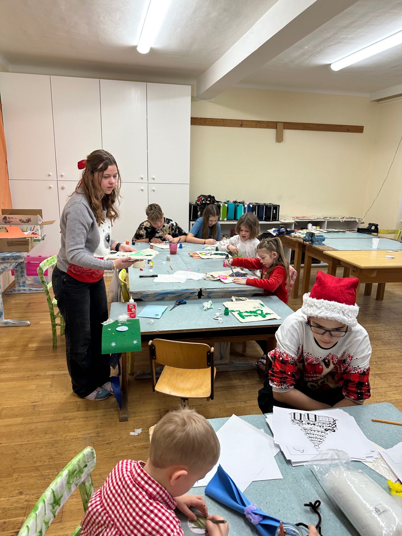 Ein Kunstunterricht mit Kindern, eine Frau in einem Pullover leitet, Kinder in festlichen Mützen malen, Holzstühle und Tische in einem Raum mit weißen Schränken und einem Holzboden.