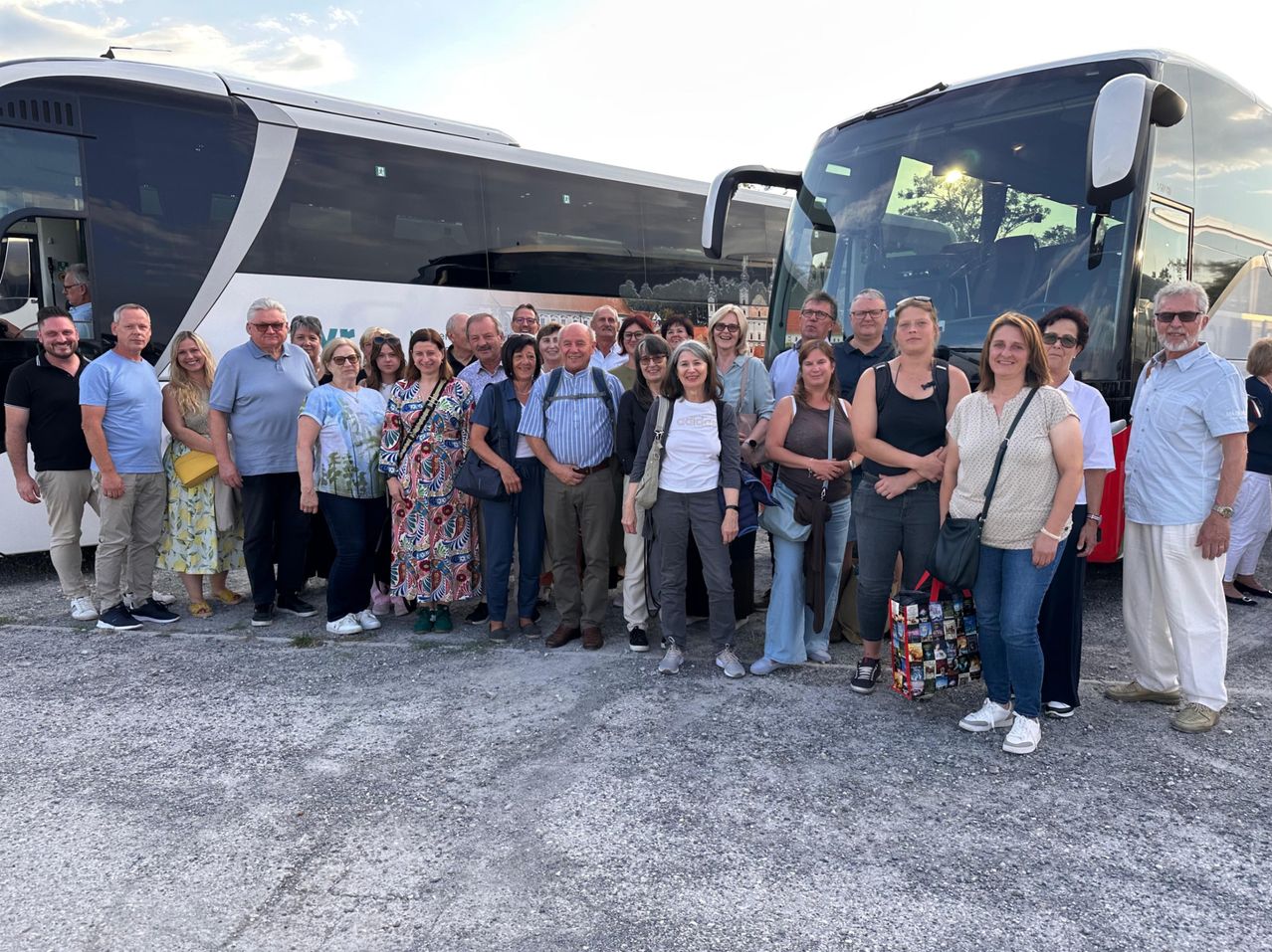 Eine Gruppe von Menschen, wahrscheinlich Touristen, steht vor zwei Bussen auf einer Kiesoberfläche. Einige tragen Sonnenbrillen und haben Taschen bei sich.
