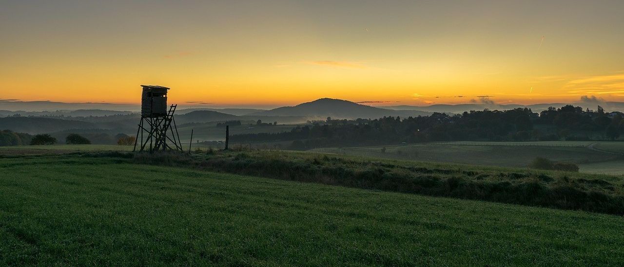 Ein ruhiges Feld mit einem Jagdturm bei Sonnenuntergang, umgeben von einem Zaun und Bergen in der Ferne.