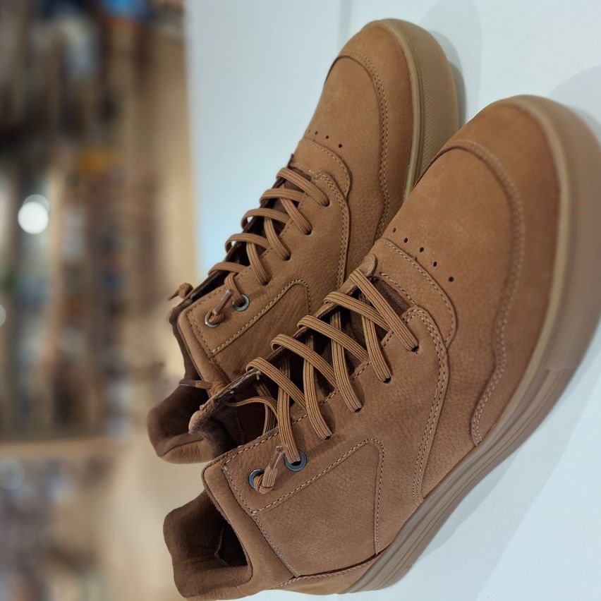 Ein Paar braune Leder-High-Top-Sneaker mit Schnürsenkeln und blauen Ösen hängen an einer weißen Wand. Die Schuhe haben eine dicke Sohle und ein strukturiertes Lederobermaterial.