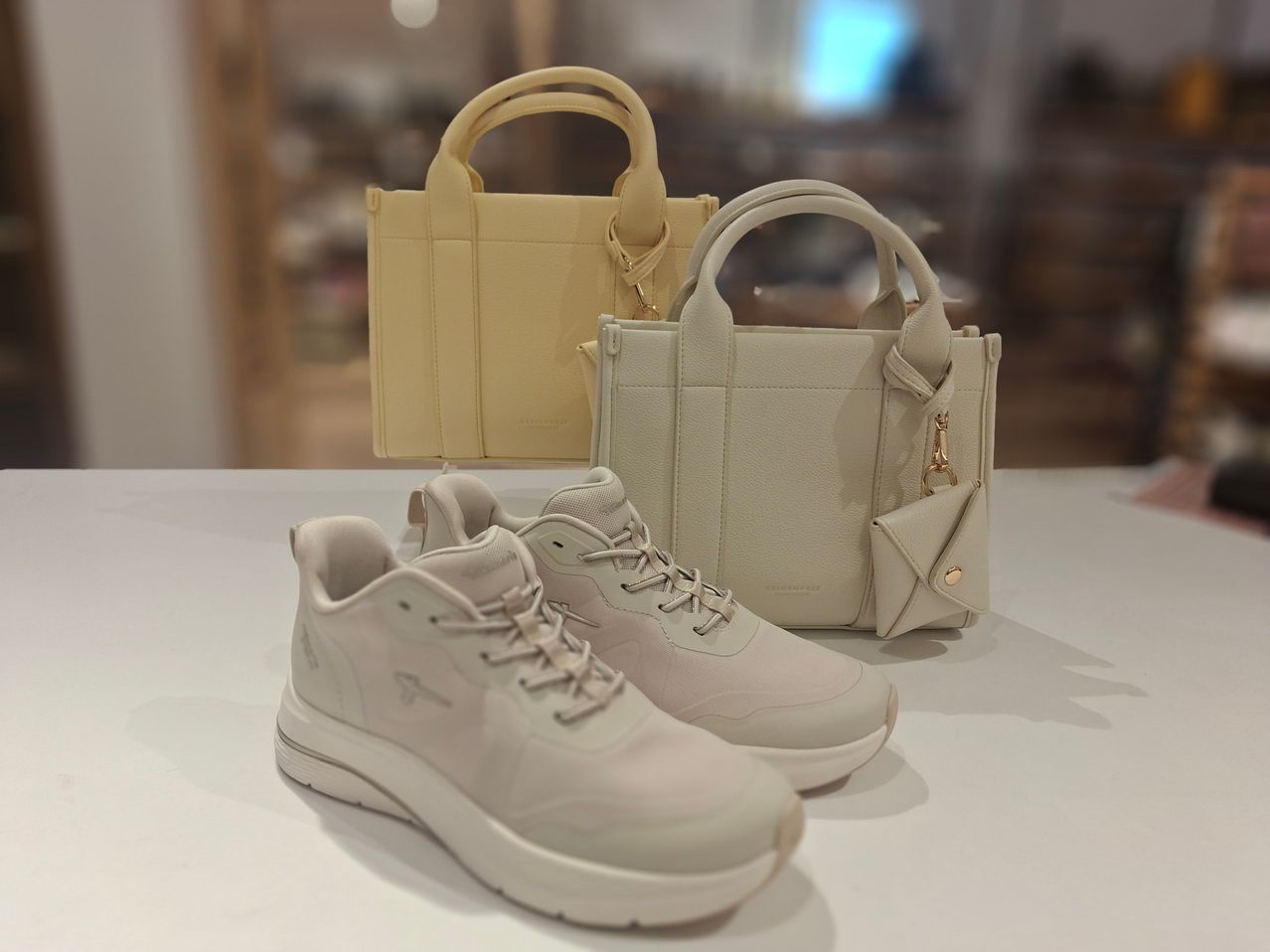 Two pairs of white sneakers with laces and soles are on a white table, with a beige handbag and a small beige wallet behind them.