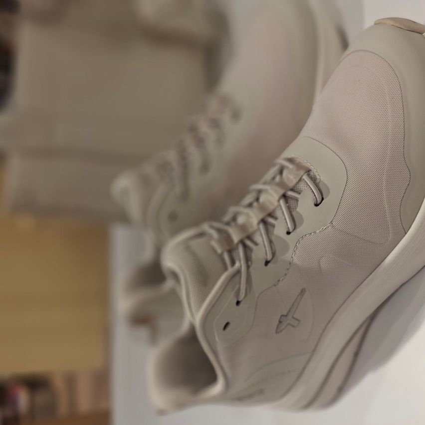 A pair of light beige sneakers with a mesh design, laces, and a subtle logo on the side, displayed on a white surface.