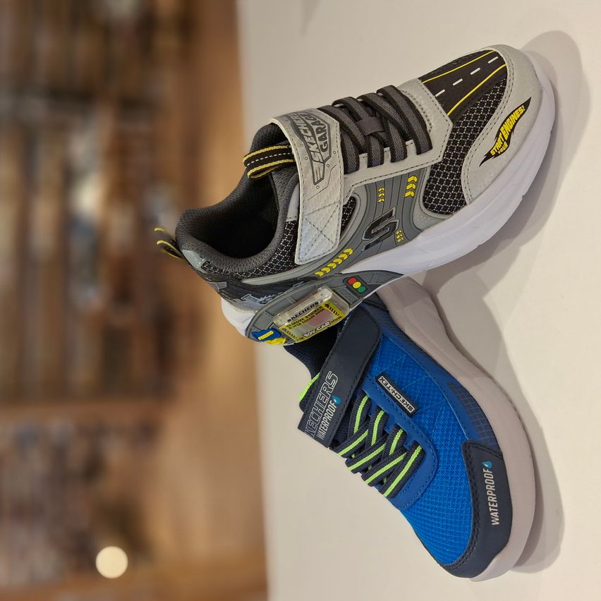 Zwei Paar Schuhe sind ausgestellt, eines grau und schwarz, das andere blau und grn, an einer weien Wand hngender.