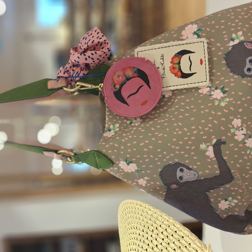 Eine grüne Handtasche mit rosa und schwarzen Blumendesigns, einer Affenillustration und einem rosa Etikett mit einem Frauengesicht. Sie hat eine rosa Schleife, grüne Riemen und eine runde rosa Geldbörse.