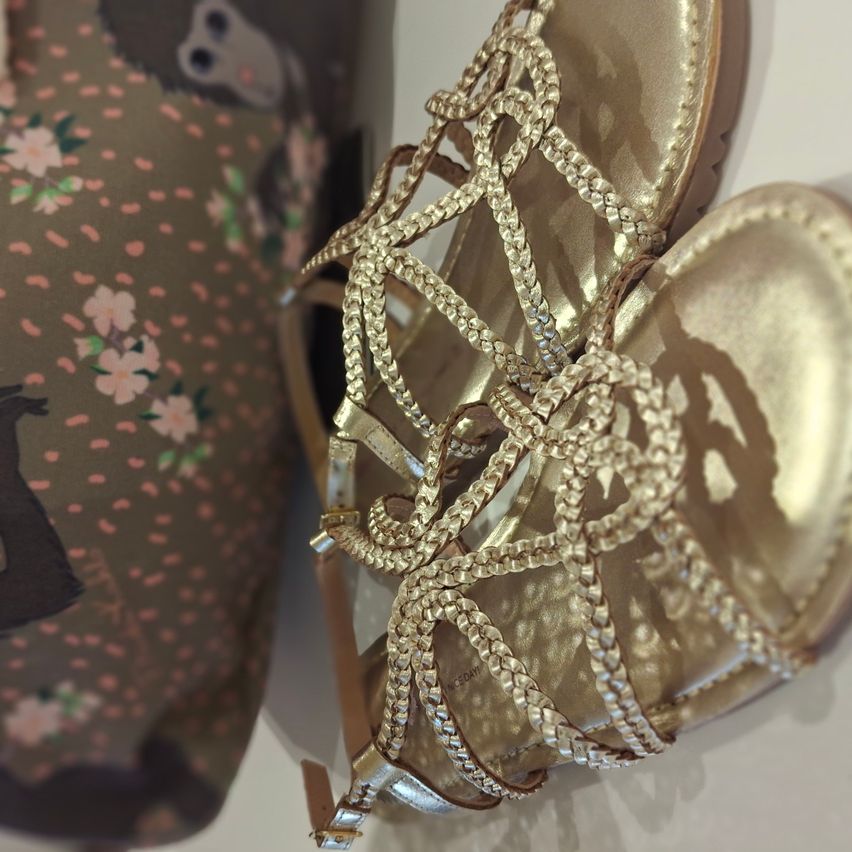 Goldene Sandalen mit aufwendigen Kettenglieder-Riemen und glänzenden Verzierungen, vor einem floralen Hintergrund präsentiert.