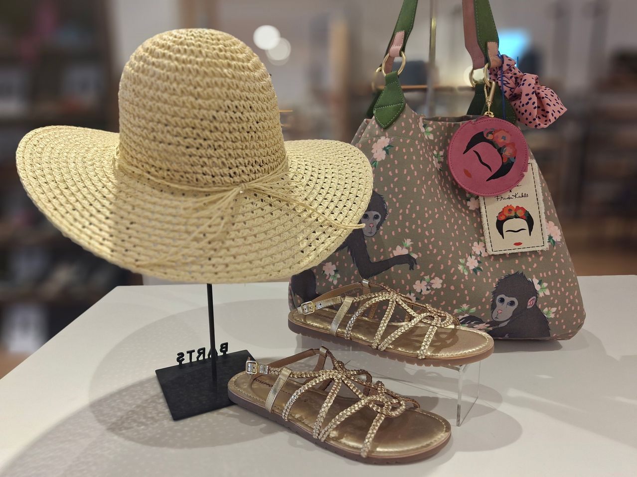 Ein Strohhut, Sandalen und eine Handtasche mit Frida-Kahlo-Design sind auf einem weißen Tisch ausgestellt. Der Hut ist braun und die Sandalen golden. Die Handtasche hat ein Affendesign.