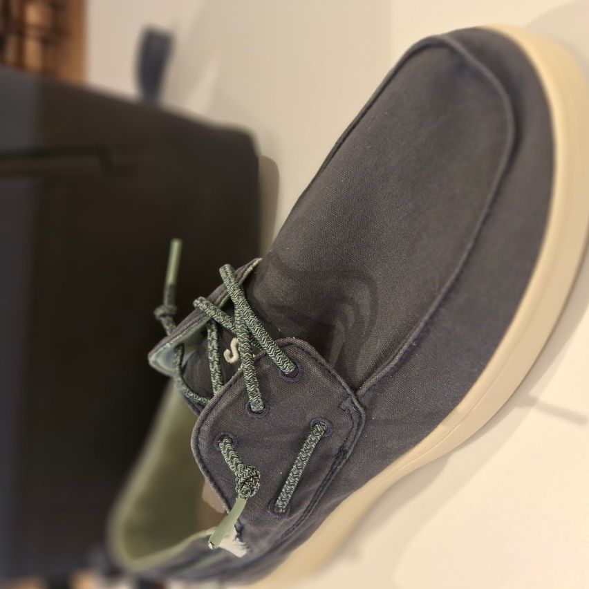 Ein Nahaufnahme eines grauen Wildlederschuhs mit beigefarbener Sohle, der Schuh hat grüne Schnürsenkel.