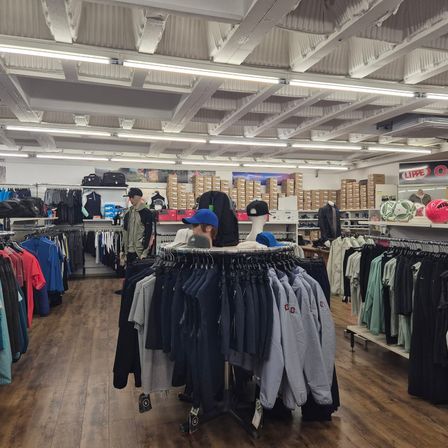Ein Bekleidungsgeschäft mit einer Vielzahl von Kleidungsstücken an Kleiderbügeln und Regalen. Auch Mannequins und Taschen sind ausgestellt. Die Decke hat mehrere Lichter.