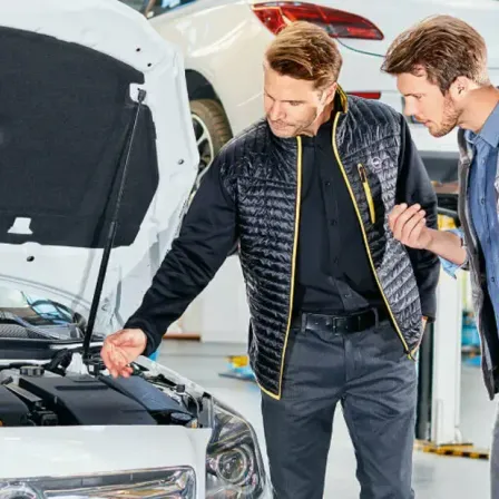 Zwei Männer in einer Garage, einer zeigt auf die offene Motorhaube eines Autos, während der andere zusieht.