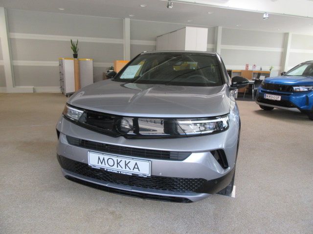 A silver car with the license plate 'MOKKA' is parked in a showroom with a blue car in the background.