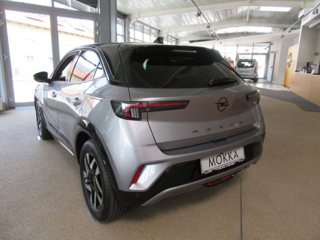 The rear end of a silver Opel Mokka is displayed in a showroom with large windows and a white ceiling. The car has a sleek design, with a license plate reading 'MOKKA'.