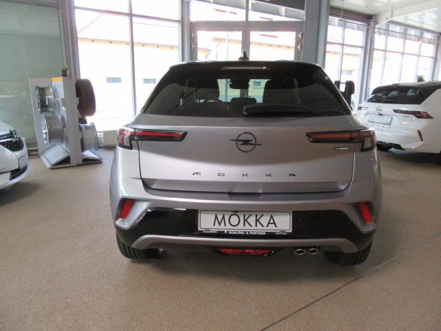 The rear view of a silver Opel Mokka car parked inside a showroom with other cars. The car has a license plate that reads 'MOKKA'.