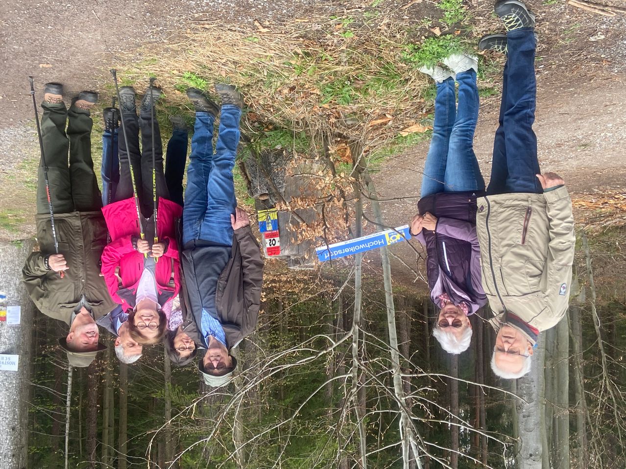 Six individuals, wearing jackets and holding sticks, are standing in a forest, smiling for a photo. A sign reading 'Horche/Hoerchele' and another with '20' is visible in the background.