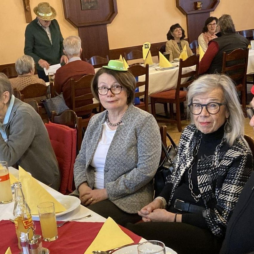 Mehrere ältere Personen sitzen an Tischen in einem Restaurant. Eine Frau trägt einen grünen Hut mit einem gelben Bogen. Eine andere Frau trägt eine Brille und eine Halskette.